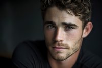 Close-up portrait of young man with curly hair and light stubble against dark background, conveying thoughtful expression and natural lighting.