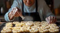 Senior woman in black apron sprinkling sugar on freshly baked butter cookies cooling on wire rack, close up view of hands dusting traditional homemade pastry.