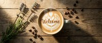 Latte art coffee cup with Welcome text and heart pattern on rustic wooden table, top view with lavender and scattered beans.