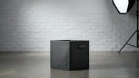 Modern black smart speaker cube on wooden floor against white brick wall in minimalist studio setting with professional lighting equipment.