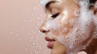 Side profile of young woman with glowing skin and water droplets splashing on face against pink background, showcasing skincare and beauty concept.
