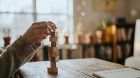 Person stacking wooden blocks on table in cozy cafe setting with warm bokeh lights creating peaceful atmosphere for mindfulness and focus activities.