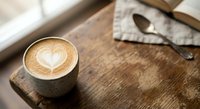 Coffee cup with heart latte art on rustic wooden table next to open book and spoon. Cozy cafe atmosphere for morning routine and relaxation concepts.