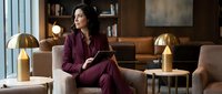 Confident Middle Eastern businesswoman in burgundy suit holding tablet, sitting in modern luxury office lounge with gold lamps, thoughtful expression.