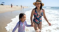 Happy Asian mother and daughter enjoying summer beach vacation together, walking hand in hand along sandy shoreline with ocean waves and blue sky.