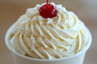 Creamy vanilla soft serve ice cream swirled in white paper cup topped with bright red maraschino cherry, close up view showing smooth texture and detailed piping pattern.