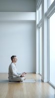 Middle-aged Caucasian man in white clothing practicing meditation in modern minimalist room with large windows and natural light on yoga mat.