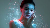 Young Asian woman with glowing makeup and face art illuminated by neon red and blue lights against dark background with magical sparkle effects and light particles.