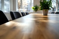 Modern wooden conference table surface with selective focus and natural lighting effect, black chairs and potted plant in bright office setting with windows.
