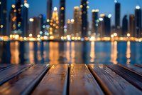 Wooden deck planks in foreground with defocused city skyline and water reflections at night, illuminated urban landscape with bokeh lights creating dreamy atmosphere.