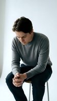 Young Caucasian man in gray sweater sitting on chair looking down with contemplative expression showing stress, anxiety or depression in minimalist setting.