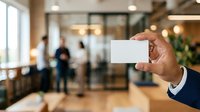Businessman holding blank white business card in modern office lobby with colleagues and glass doors in background, suitable for corporate branding and professional networking concepts.