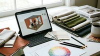 Interior designer workspace with tablet showing 3D room visualization, fabric samples, color wheel charts and design materials on desk near window.