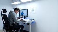 Young Caucasian businessman in gray hoodie attending virtual meeting on computer in modern white office workspace for remote work and teleconferencing.