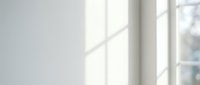 Bright natural light streaming through window panes casting geometric shadows on white wall, creating minimalist interior ambiance suitable for backgrounds and design projects.