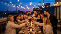Group of diverse young adults toasting with wine glasses at outdoor rooftop dinner party under string lights during twilight hour with city skyline background.