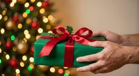 Elderly hands holding wrapped Christmas gift with red ribbon against blurred holiday tree lights background for seasonal giving and celebration.