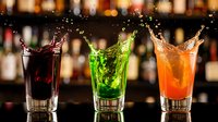 Three colorful cocktails splashing in glasses at bar counter with blurred bottle background. Dynamic liquid motion in red, green and orange drinks.