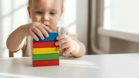 Toddler child playing with colorful wooden building blocks stacking toys on white table developing fine motor skills and creativity in bright room.
