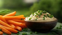 Fresh carrot sticks served with creamy herb dip garnished with chopped chives in rustic brown bowl against blurred green background, appetizing healthy snack.