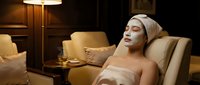 Young woman relaxing in luxury spa chair with facial mask and towel wrap enjoying wellness treatment in elegant salon interior.