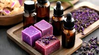 Handmade purple soap bars with essential oil bottles and dried lavender flowers on wooden board, natural spa wellness composition in warm lighting.