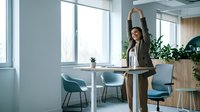Young professional businesswoman stretching arms up during break in modern office conference room with natural light and plants.