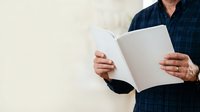 Middle-aged man with medium complexion wearing dark blue plaid shirt holding open blank white document or brochure against neutral background for business, marketing, or corporate communications.