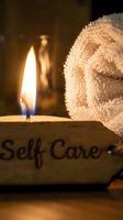 Lit candle beside a rolled white towel and a stone sign reading Self Care, creating a warm, relaxing spa atmosphere for wellness concepts.