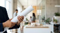 Businessman holding rolled white blueprint in modern office corridor with colleagues and plants in background, representing architecture project planning and professional construction management.