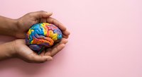 Hands gently protecting colorful brain model on pink background representing mental health care, psychology support and neurological wellness concept.