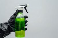 Hand in black protective glove holding green cleaning spray bottle against gray background for household maintenance and sanitation tasks.
