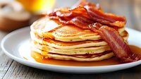 Stack of golden pancakes topped with crispy bacon strips and maple syrup on white plate, breakfast comfort food for restaurant menu design.