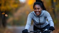 Young African American woman laughing while riding bicycle in autumn park, wearing grey sports jacket. Natural fall foliage and bokeh background creates joyful outdoor scene.