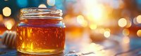 Golden honey jar glowing in warm sunlight with bokeh lights background, wooden dipper on jar rim creates natural organic food composition.