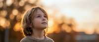 Young child with curly hair looking up thoughtfully during golden hour sunset with warm bokeh lights creating dreamy atmospheric portrait for family content.