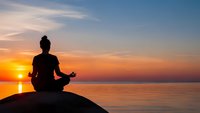 Woman silhouette meditating in lotus pose on rock by calm ocean at sunrise. Peaceful yoga practice with golden sky reflection on water for wellness.