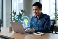 Asian businessman working on laptop with digital innovation icons and lightbulb hologram in modern office environment for technology and business concepts.