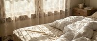 Cozy unmade bed with white linens in warm sunlit bedroom with lace curtains and wooden furniture creating peaceful morning atmosphere.