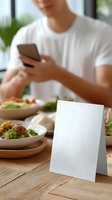 Young Asian man in white t-shirt using smartphone during lunch with blank menu card on wooden table. Restaurant setting with fresh salads and dishes.