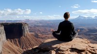 Young woman in black clothing meditating on cliff edge overlooking dramatic red rock canyon landscape with mountain peaks and blue sky in background. Peaceful mindfulness moment.