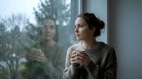 Thoughtful young woman with dark hair in casual sweater holding warm beverage, gazing through rainy window at forest landscape, contemplative mood, suitable for wellness and lifestyle content.