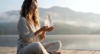 Young woman practicing mindfulness meditation with crystal ball at serene mountain lake during golden hour sunrise, promoting wellness and spiritual healing.