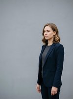 Young woman with shoulder-length wavy blonde hair and fair complexion wearing navy blazer over gray top, standing against neutral gray background. Corporate and professional styling suitable for business, corporate portraits, and professional headshots.