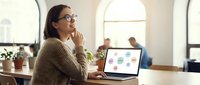 Woman with glasses working on laptop in bright office space, analyzing data charts and graphs for business planning and professional development.
