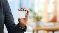 Businessman with dark skin tone holding blank white business card in office environment with blurred background for corporate branding and contact information display.