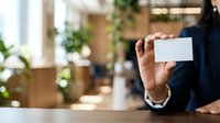 Businessman in dark suit holding blank white business card in modern office with plants and natural light.