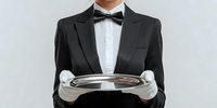 Professional waiter in formal black tuxedo and bow tie holding empty silver serving tray with white gloves against neutral background.