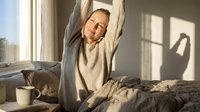 Woman stretching in bed after waking up, wearing cozy sweater, morning sunlight streaming through window, relaxed lifestyle moment.