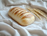 Fresh baked white bread loaf with golden crust and diagonal scoring marks on white linen cloth, accompanied by wheat stalks, food photography.